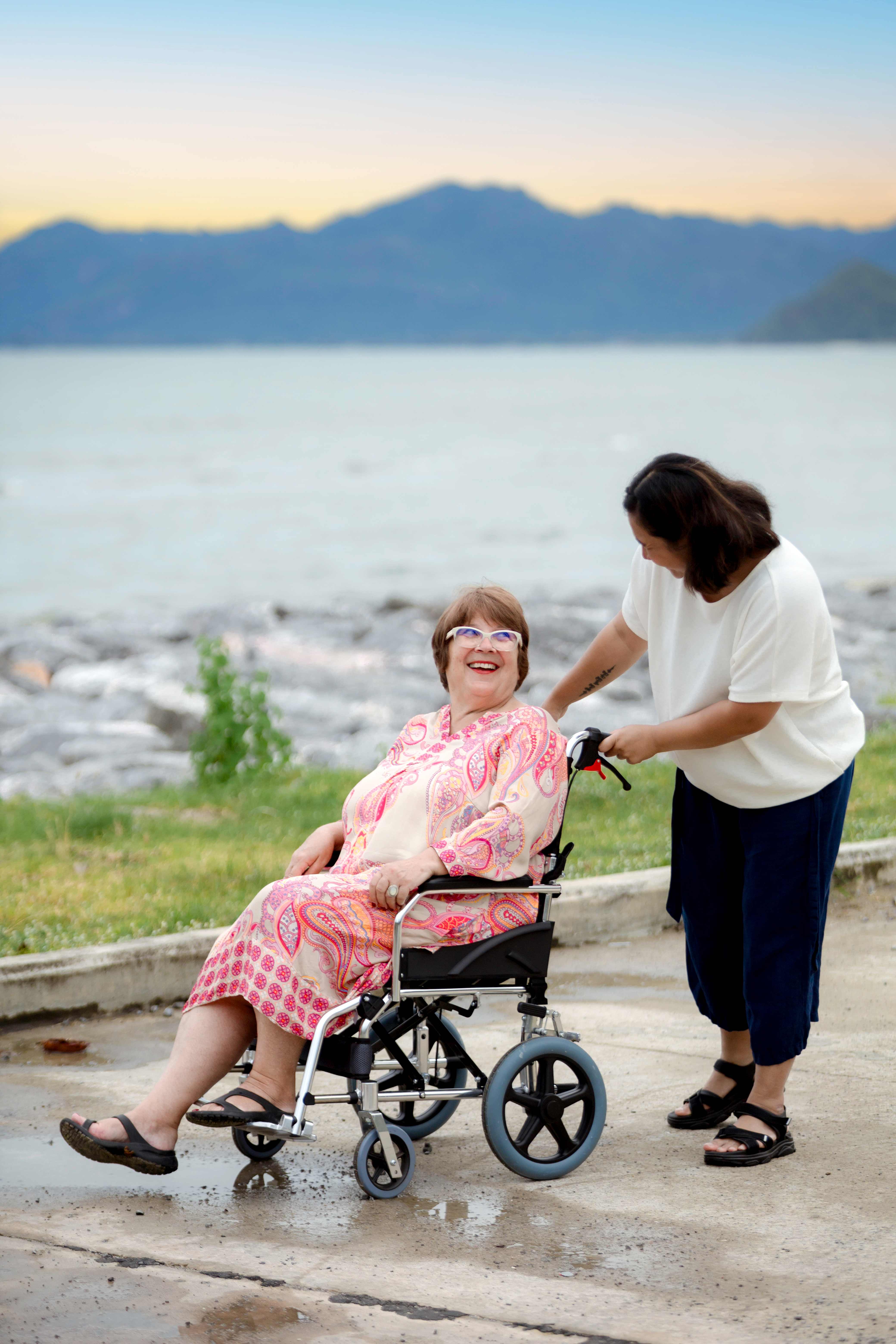 Lachende Seniorin im Rollstuhl mit Betreuerin am Meer bei Sonnenuntergang vor Bergpanorama